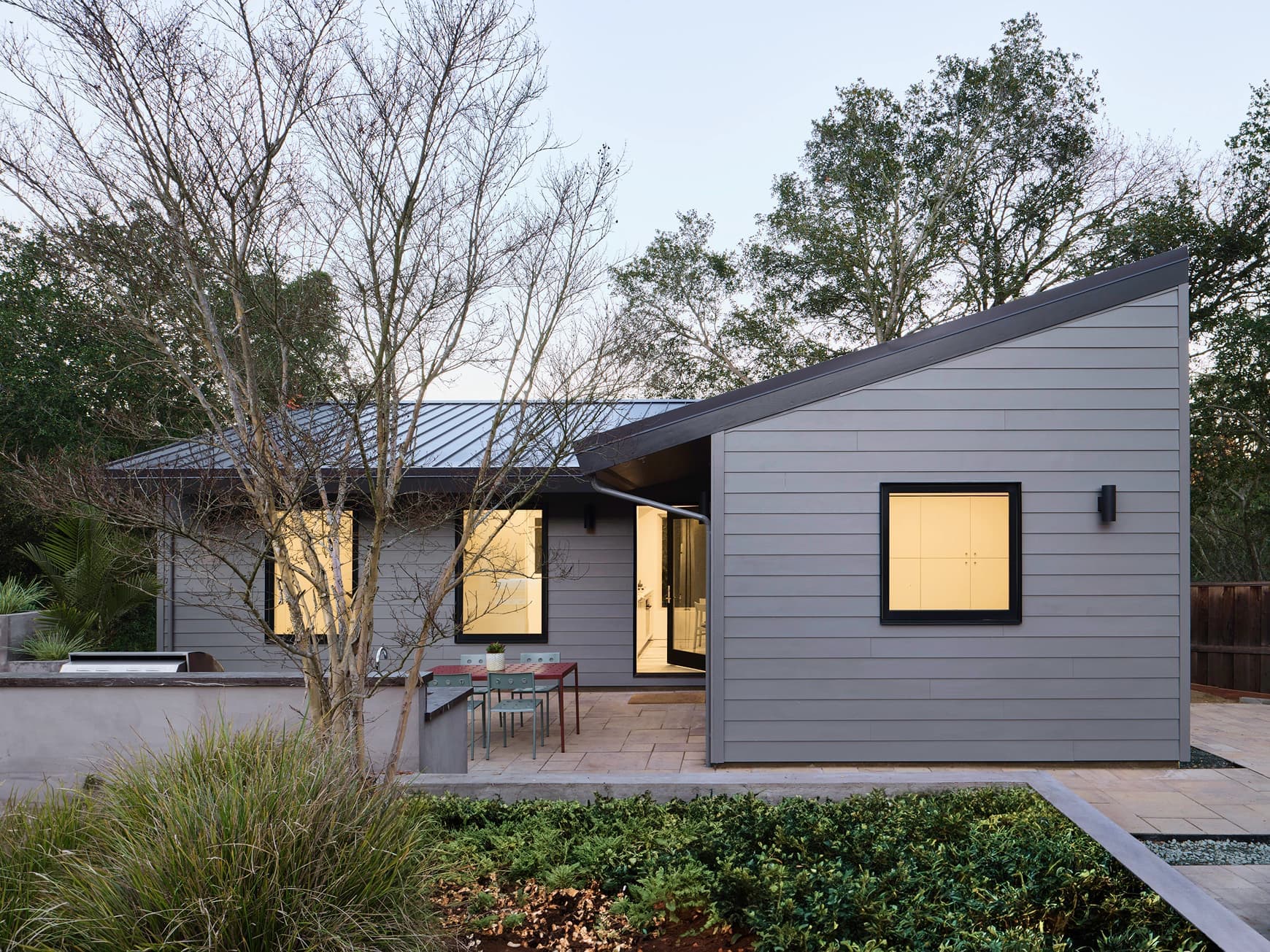 A finished single-story gray home with a shed roof and lit windows, behind a patio and garden
