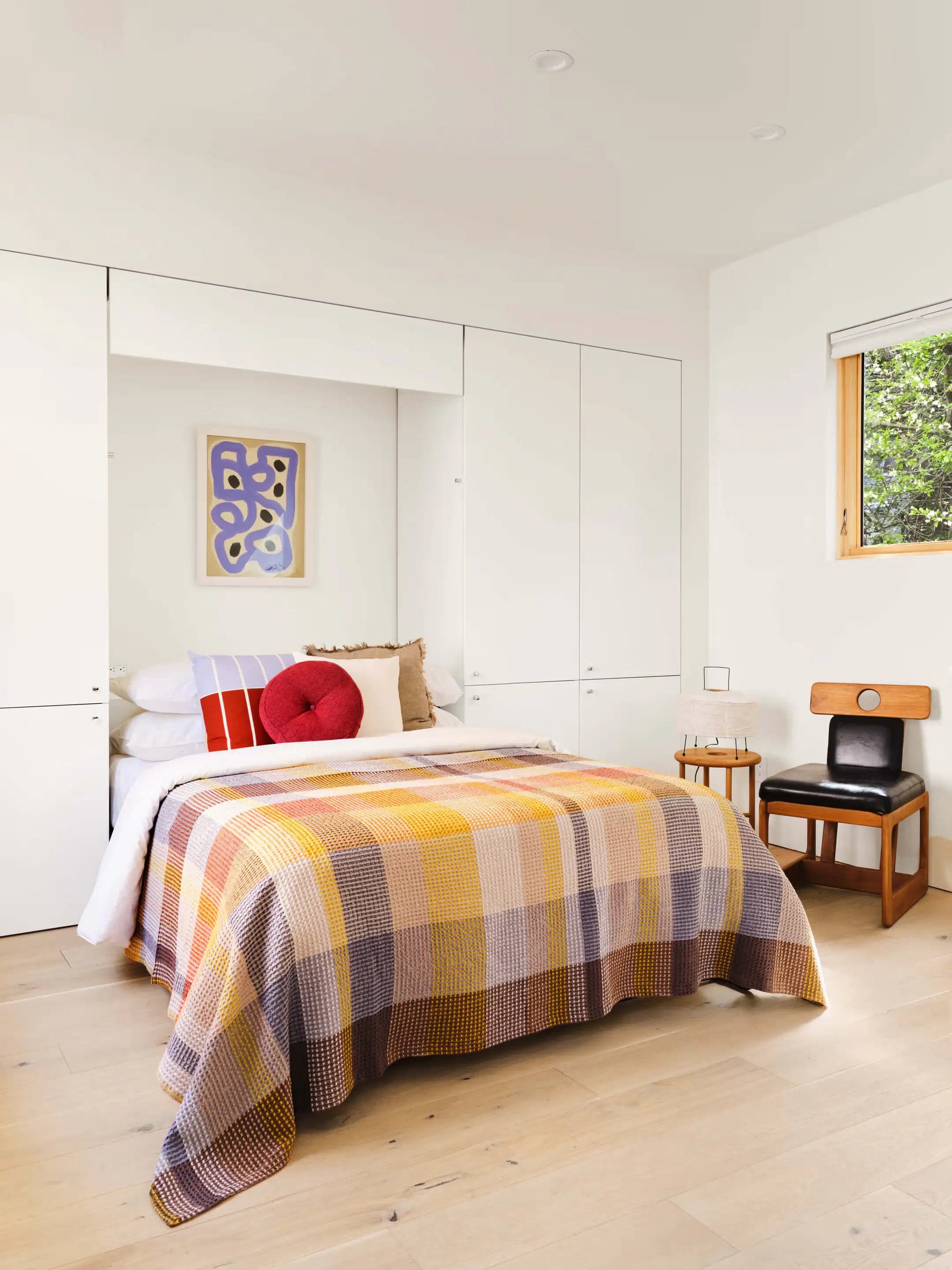 A bedroom with white built-in cabinets around the bed, a plaid blanket, and a wood and leather chair by the window