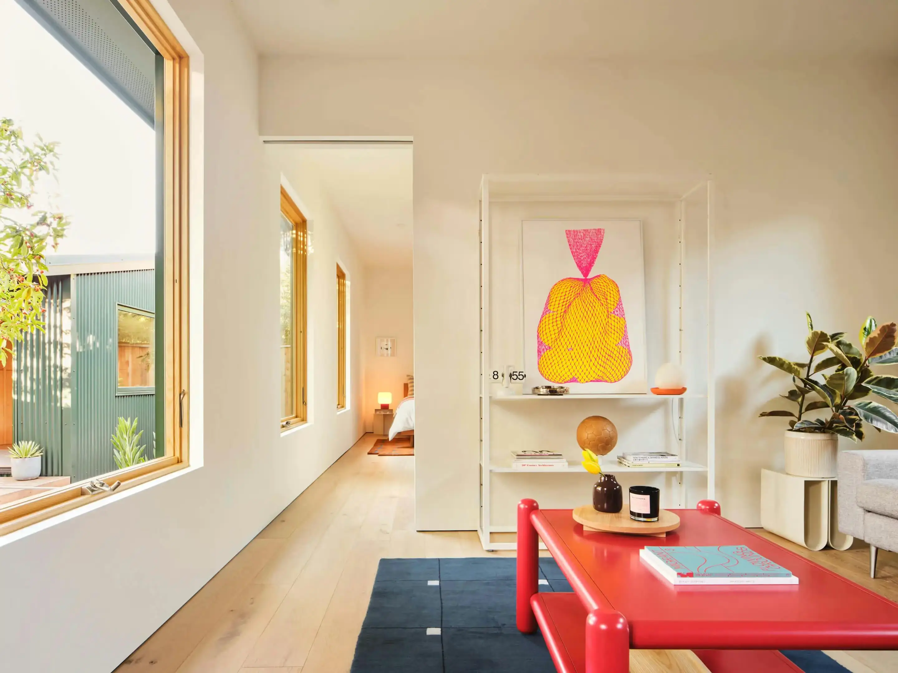 A living room with a red coffee table and an art print on an open shelf, beside a hallway of windows leading to a bedroom