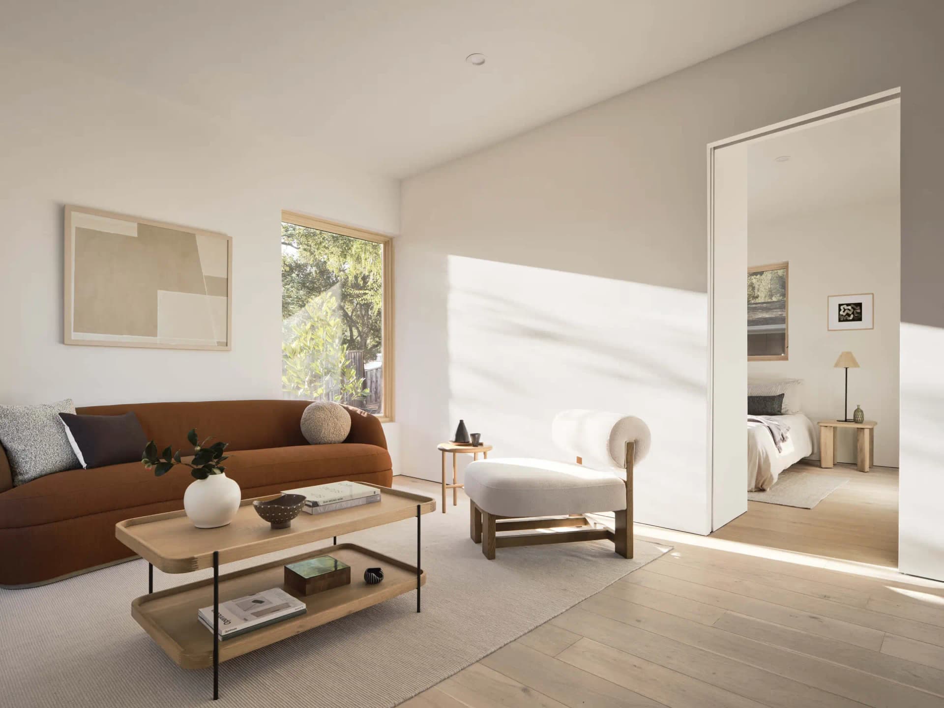 A sunlit living room with a curved rust-colored sofa, an oak coffee table, and a doorway to a bedroom